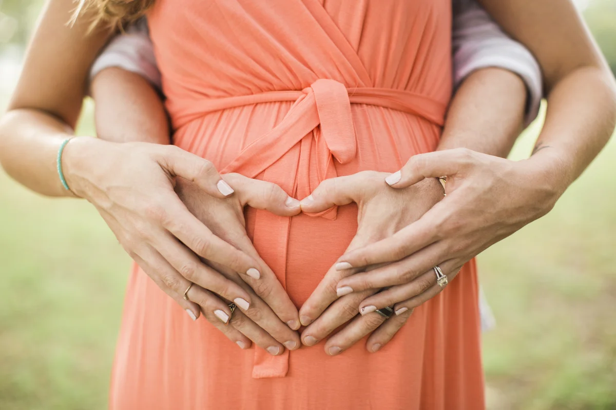 closeup of a pregnan womns midsection wearing a coral dress, her husband hugging her from behind forming a heart on their baby bump